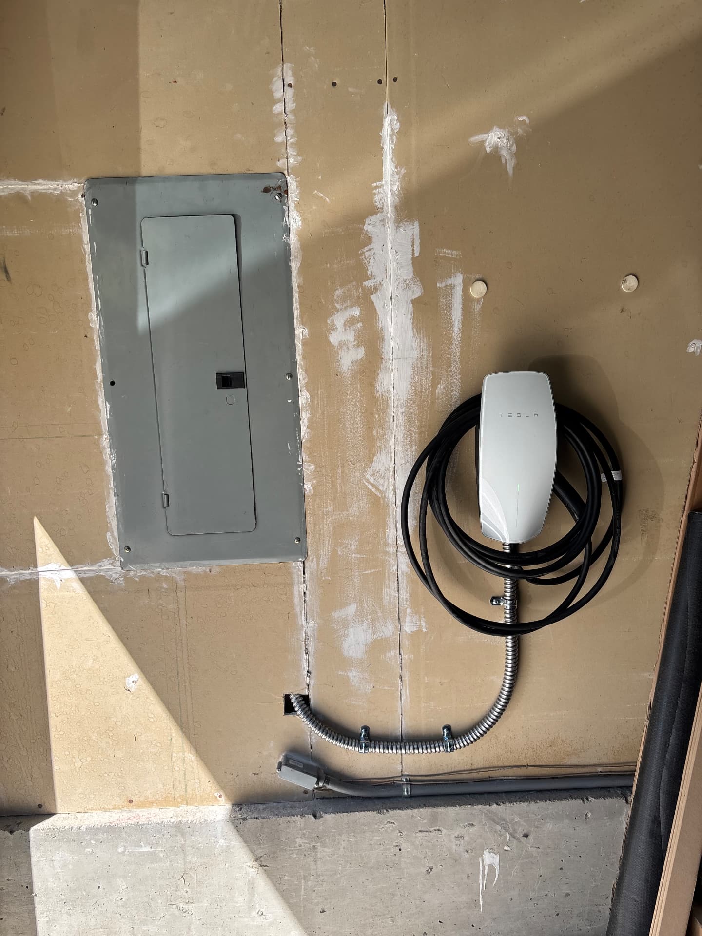 Tesla Wall Connector beside a garage electrical panel with flexible metal conduit